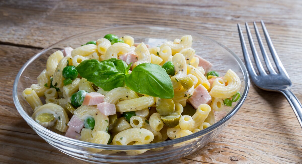 Durchsichtige Schale gefüllt mit Nudelsalat mit Fleischwurst und Erbsen. 
