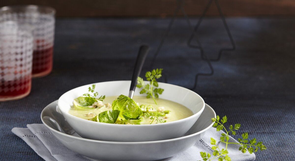Die Rosenkohl-Cremesuppe in weißem Geschirr auf einem dunkelblauen Tisch und dunkelblauer Wand. Alles von der Seite fotografiert.