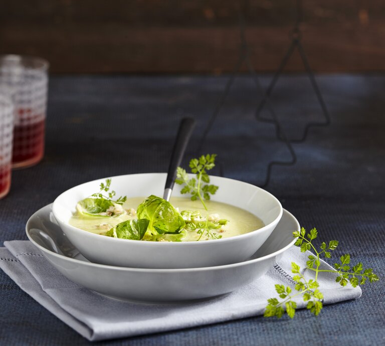 Die Rosenkohl-Cremesuppe in weißem Geschirr auf einem dunkelblauen Tisch und dunkelblauer Wand. Alles von der Seite fotografiert. | © StockFood