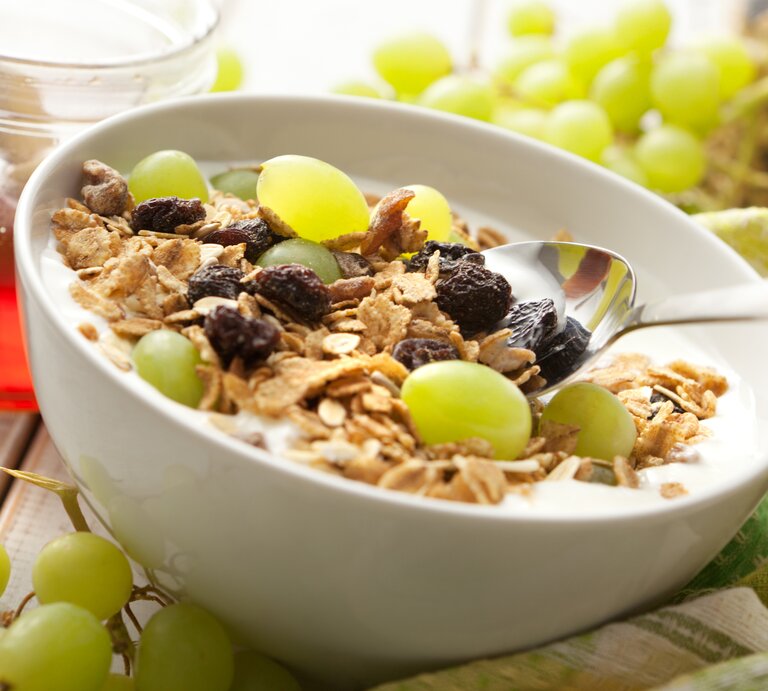 Eine weiße Schale mit dem Traubenmüsli und frischen Weintrauben auf einem dunklen Holztisch. Alles nah und von der Seite fotografiert. | © iStock/Floortje