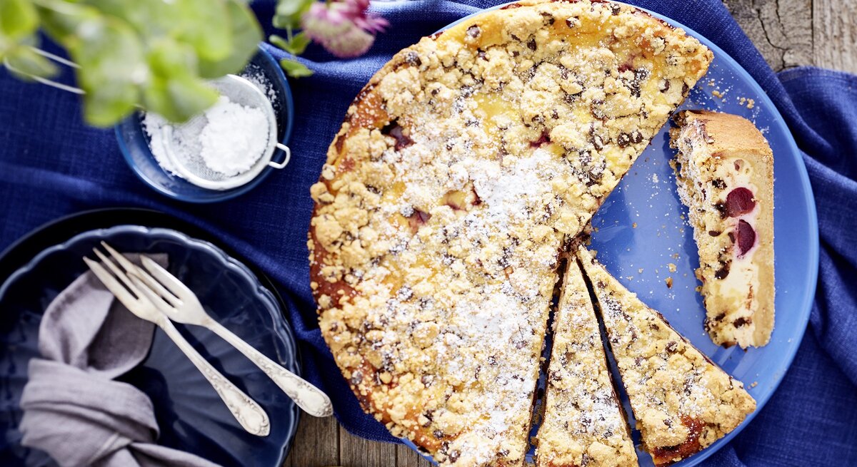 Der Käse-Kirsch-Streuselkuchen auf einem Holztisch mit blauer Tischdecke. Alles direkt von oben fotografiert.