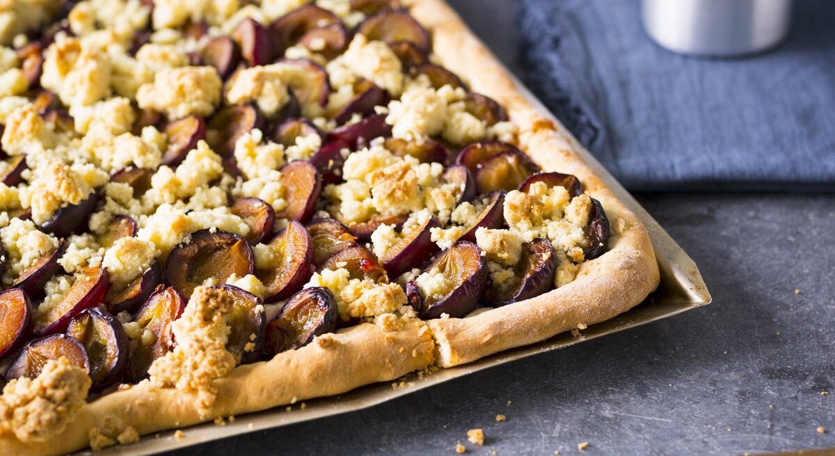 Ein silbernes Blech mit dem zuckerfreien Zwetschgenkuchen auf einem dunkelblauem Untergrund von schräg oben fotografiert.