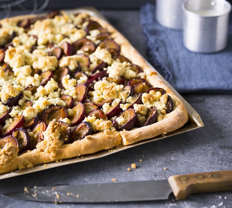 Ein silbernes Blech mit dem zuckerfreien Zwetschgenkuchen auf einem dunkelblauem Untergrund von schräg oben fotografiert. | © StockFood
