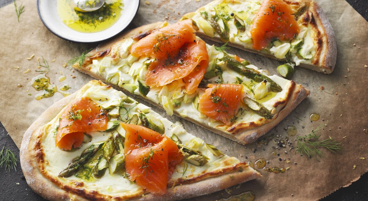 Flammkuchen mit grünem Spargel und Räucherlachs auf Backpapier und dunklem Tisch.