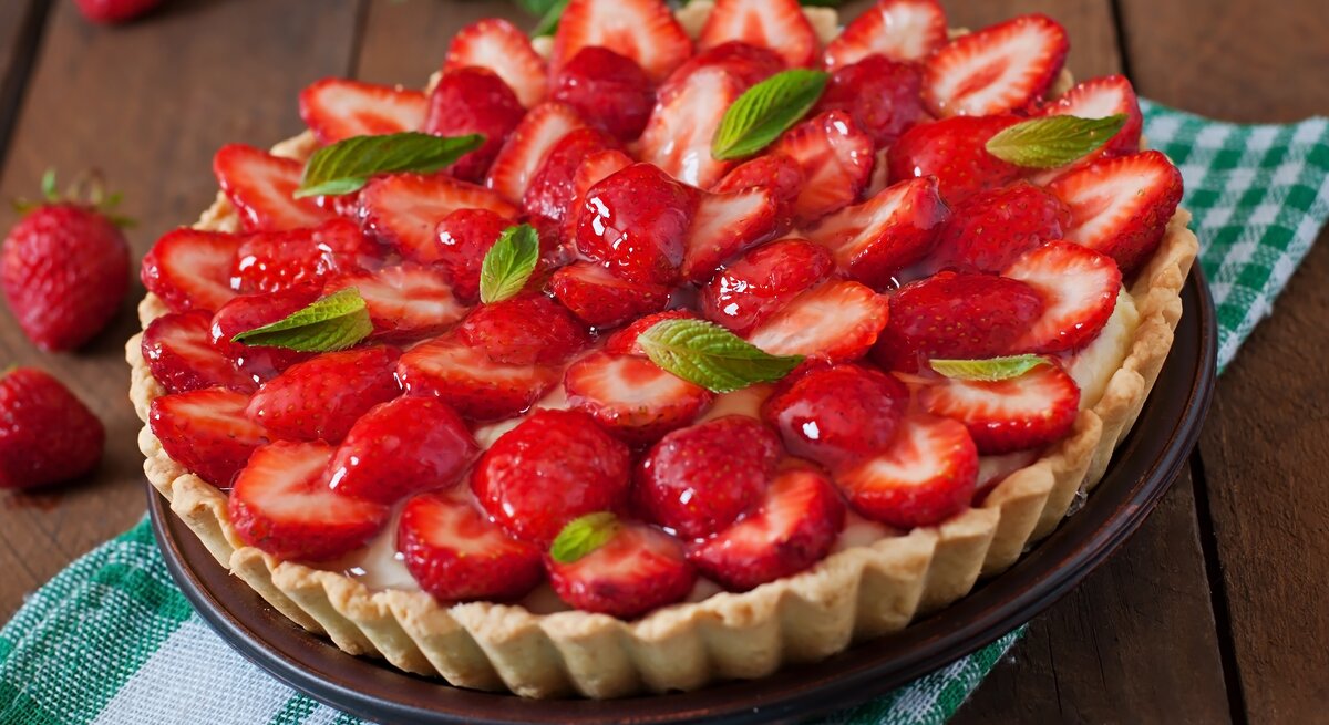 Französische Tarte mit Erdbeeren und Mascarpone-Creme auf einem grün-weiß-kariertem Tuch vor hölzernem Hintergrund, darauf Erdbeeren verteilt.