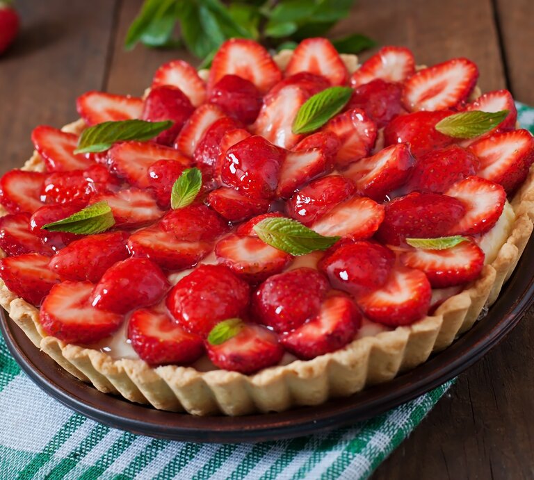 Französische Tarte mit Erdbeeren und Mascarpone-Creme auf einem grün-weiß-kariertem Tuch vor hölzernem Hintergrund, darauf Erdbeeren verteilt. | © shutterstock