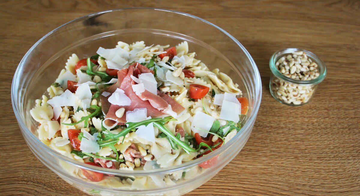 Eine Glasschüssel mit dem Nudelsalat auf einem hellen Holztisch. Alles von schräg oben fotografiert.