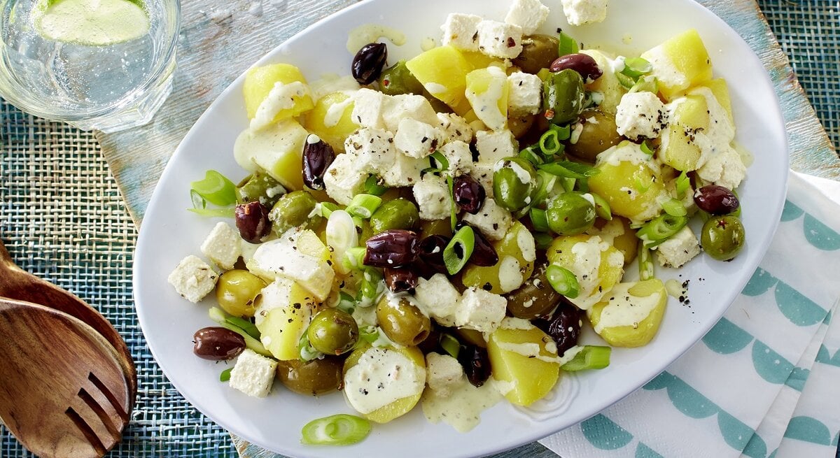 Griechischer Kartoffelsalat mit Hirtenkäse und Oliven / ovaler Teller / Salatbesteck aus Holz / Draufsicht / Nahaufnahme