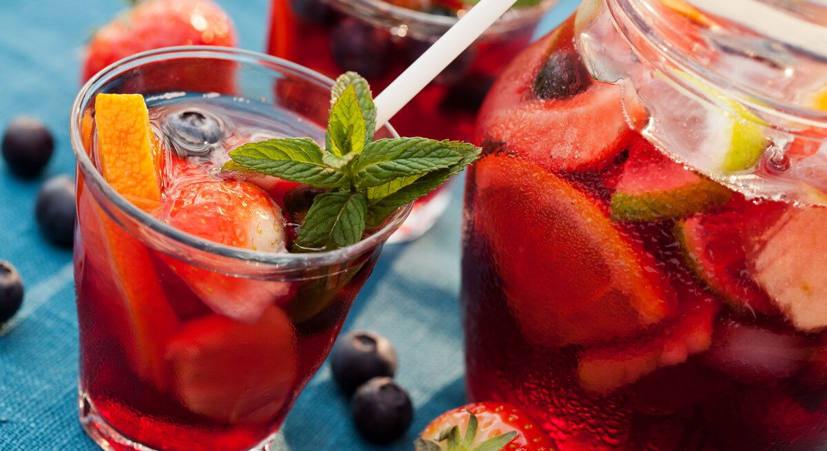 Ein paar Gläser und ein großes Glasgefäß mit Sangria und Früchten auf blauem Untergrund.