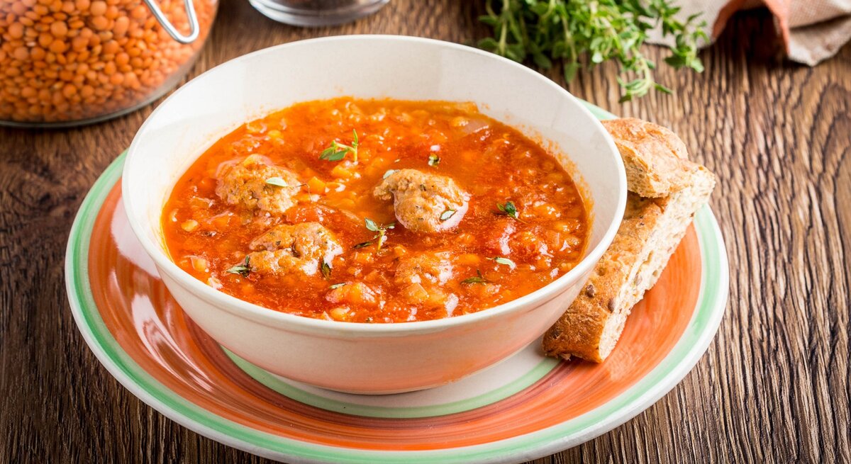 Linsen-Tomatensuppe mit Hackbällchen / runde Schüssel auf Unterteller / zwei Brotscheiben / Weckglas mit roten Linsen / Frische Kräuter / Untergrund Holz / Draufsicht, schräg