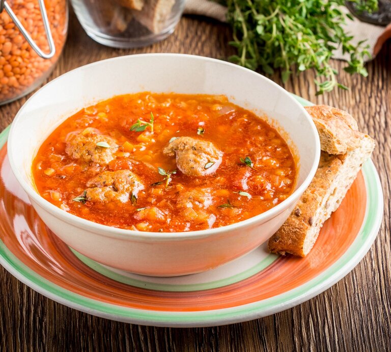 Linsen-Tomatensuppe mit Hackbällchen / runde Schüssel auf Unterteller / zwei Brotscheiben / Weckglas mit roten Linsen / Frische Kräuter / Untergrund Holz / Draufsicht, schräg | © iStock/Maria_Lapina