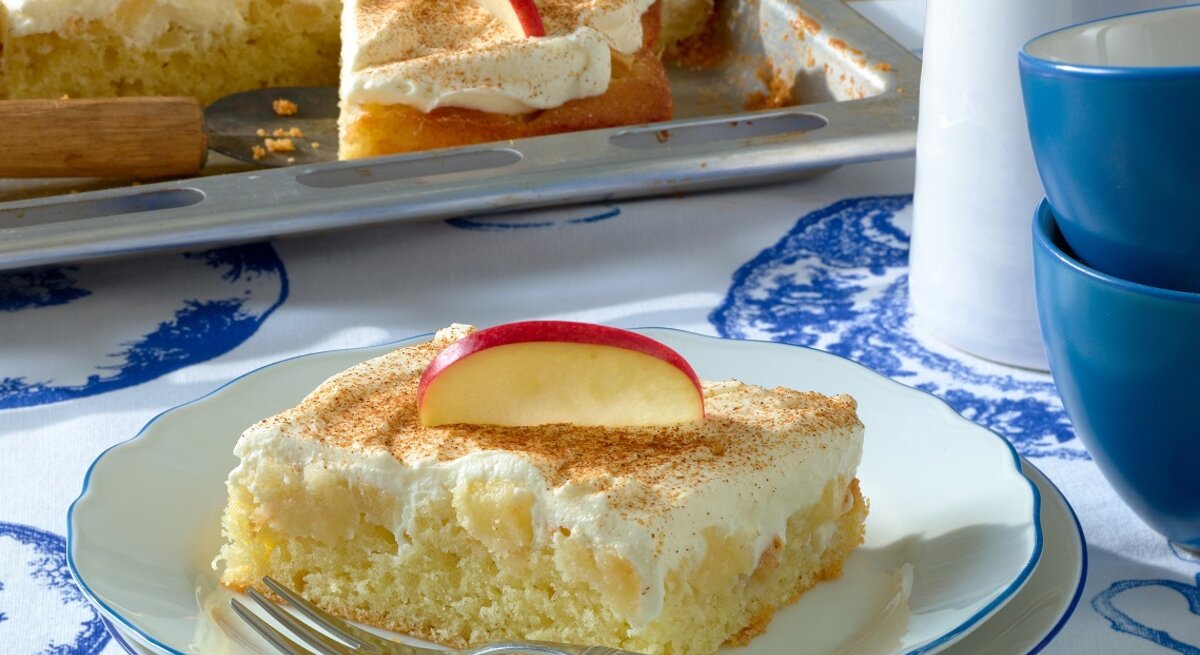 Ein Stück Apfelkuchen mit Schmandhaube auf weiß-blauem-Hintergrund. Dahinter das Kuchenblech und rechts davon Tassen.
