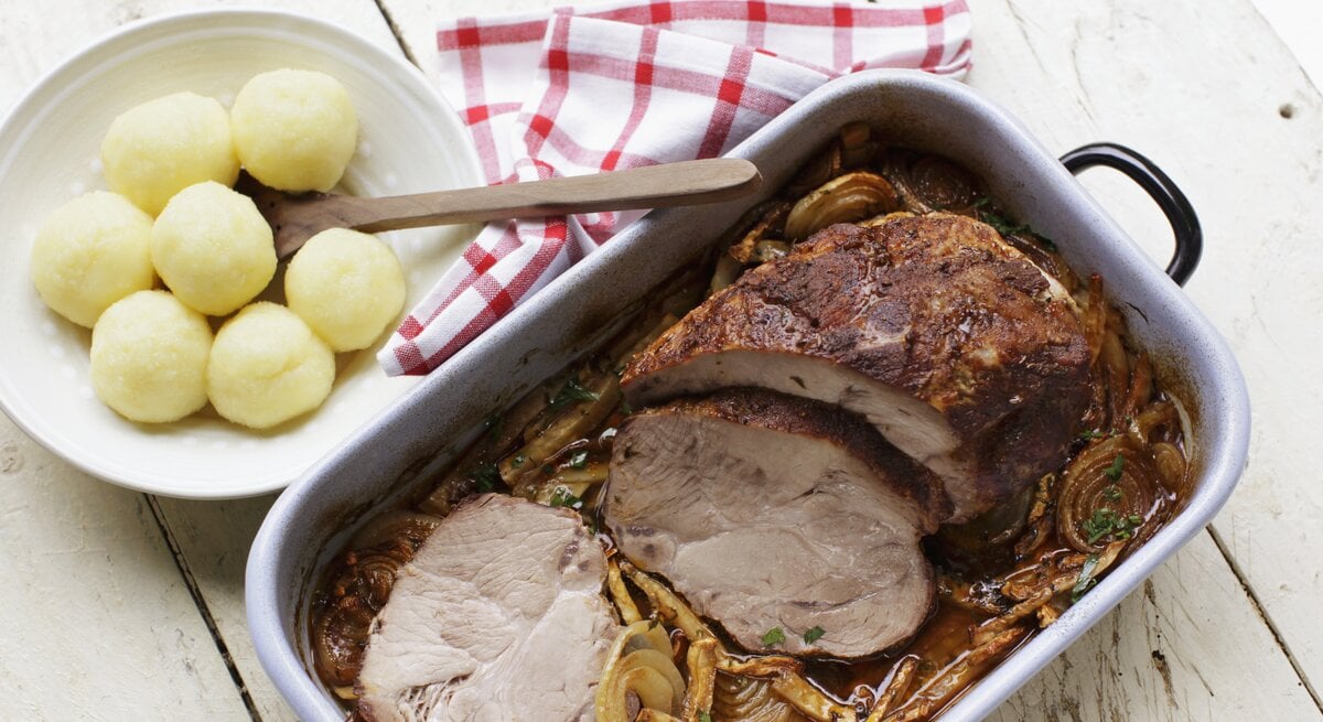 Bräter mit Schweinebraten in Soße, daneben ein Teller voll Knödel. 