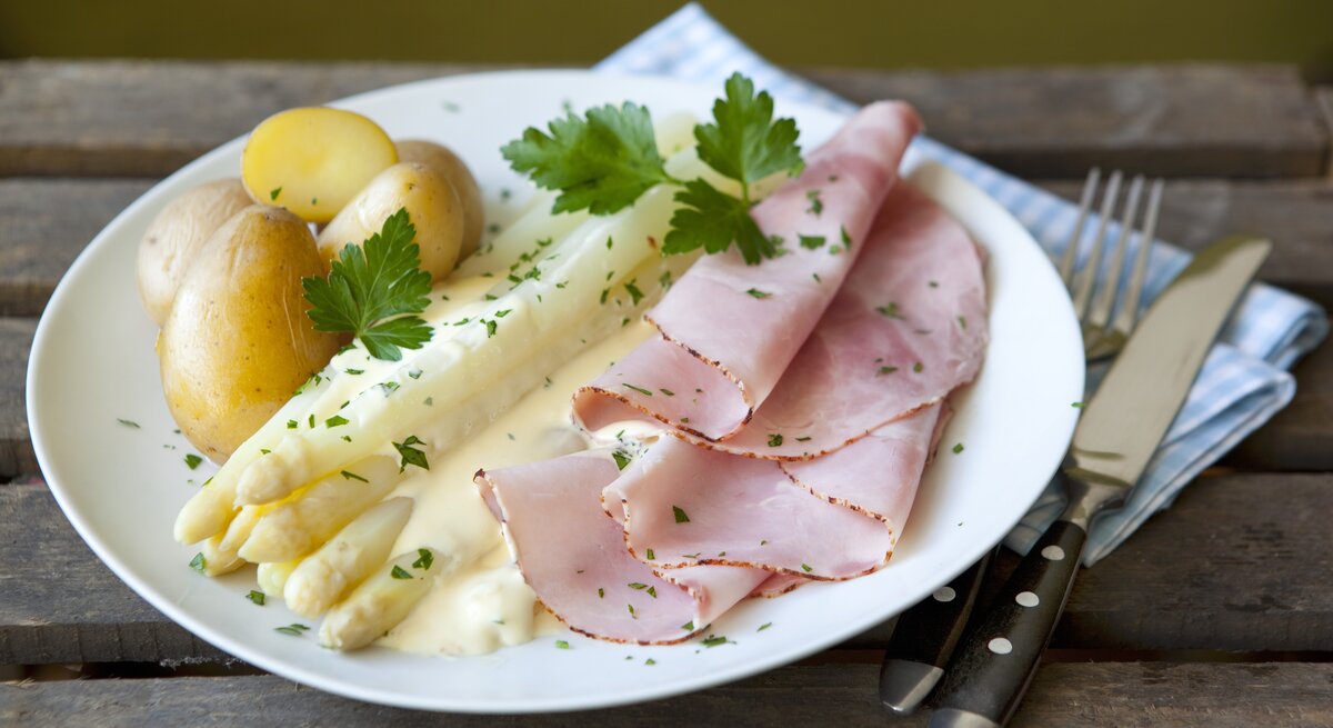 Ein weißer Teller mit Spargel, Kartoffeln, Schinken und Sauce hollandaise auf einem rustikalen Holztisch.