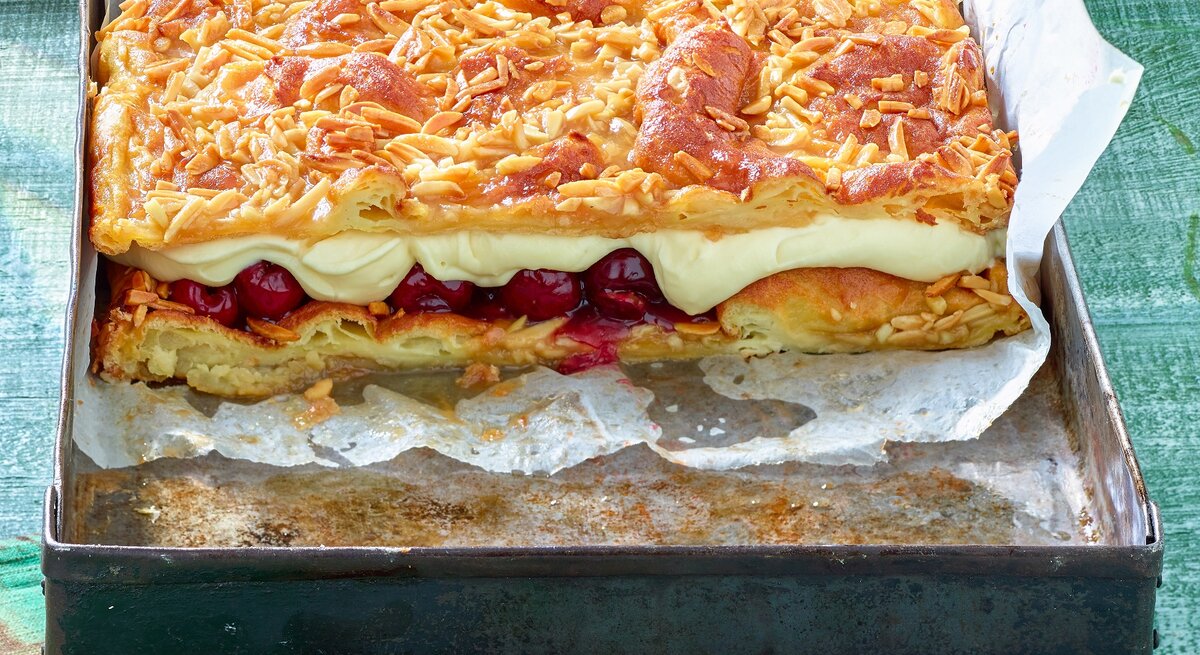 Das Bild zeigt eine Backform mit einem rechteckigen Kuchen, bestehend aus mehreren Schichten. Die oberste Schicht ist mit gerösteten Mandelsplittern bedeckt, darunter befindet sich eine Schicht aus cremiger Füllung und eine weitere Schicht mit eingelegten Früchten, vermutlich Kirschen. Darunter liegt eine Schicht aus Teig, die ebenfalls goldbraun gebacken ist.