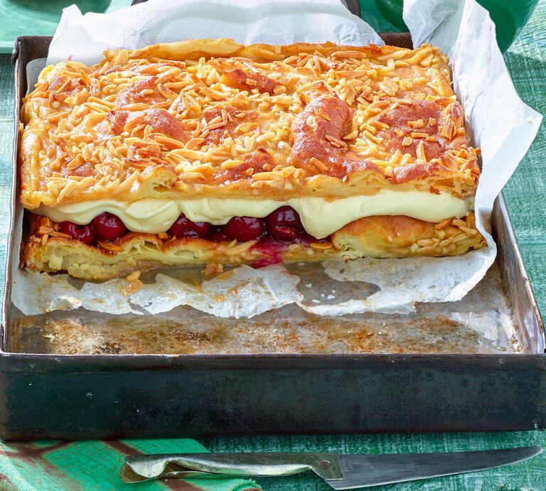 Das Bild zeigt eine Backform mit einem rechteckigen Kuchen, bestehend aus mehreren Schichten. Die oberste Schicht ist mit gerösteten Mandelsplittern bedeckt, darunter befindet sich eine Schicht aus cremiger Füllung und eine weitere Schicht mit eingelegten Früchten, vermutlich Kirschen. Darunter liegt eine Schicht aus Teig, die ebenfalls goldbraun gebacken ist. | © Matthias Liebich
