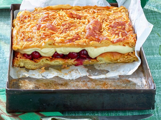 Das Bild zeigt eine Backform mit einem rechteckigen Kuchen, bestehend aus mehreren Schichten. Die oberste Schicht ist mit gerösteten Mandelsplittern bedeckt, darunter befindet sich eine Schicht aus cremiger Füllung und eine weitere Schicht mit eingelegten Früchten, vermutlich Kirschen. Darunter liegt eine Schicht aus Teig, die ebenfalls goldbraun gebacken ist. | © Matthias Liebich