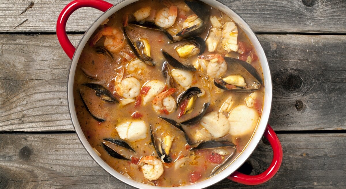 Eine köstliche Bouillabaisse mit Fisch, Muscheln, Gambas und Gemüse in einem Topf / Untergrund rustikaler Holzboden / Draufsicht