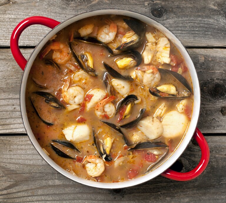 Eine köstliche Bouillabaisse mit Fisch, Muscheln, Gambas und Gemüse in einem Topf / Untergrund rustikaler Holzboden / Draufsicht | © iStock/Lara Hatarezept