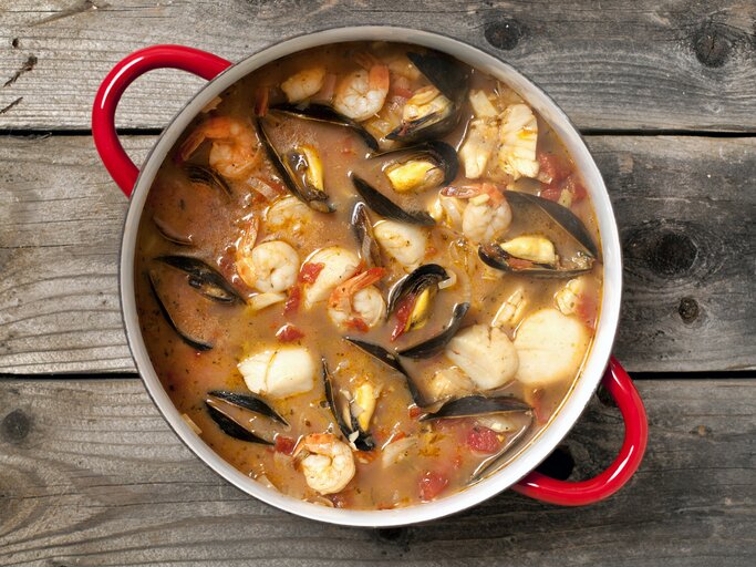 Eine köstliche Bouillabaisse mit Fisch, Muscheln, Gambas und Gemüse in einem Topf / Untergrund rustikaler Holzboden / Draufsicht | © iStock/Lara Hatarezept