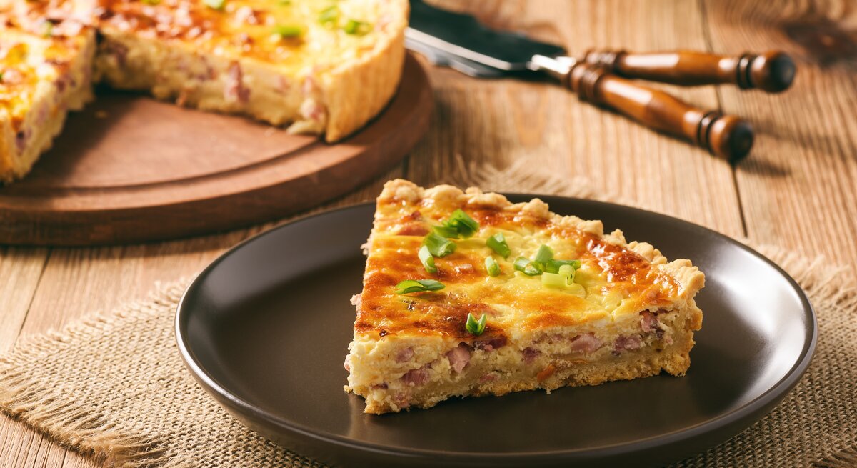  Ein Stück Quiche Lorraine auf einem dunklen Teller, garniert mit frischen Frühlingszwiebeln.