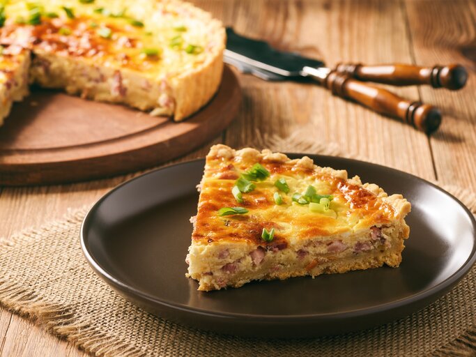  Ein Stück Quiche Lorraine auf einem dunklen Teller, garniert mit frischen Frühlingszwiebeln. | © iStock/iko636