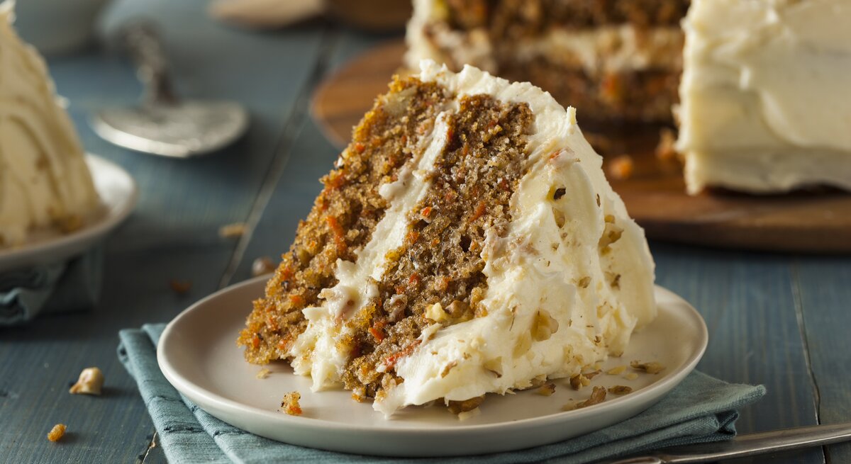 Ein Stück Karotten-Nuss-Kuchen mit Frischkäse auf  weißem Teller auf blauem Untergrund.