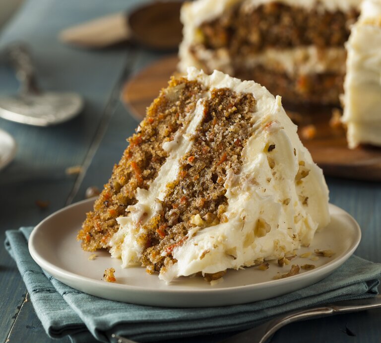 Ein Stück Karotten-Nuss-Kuchen mit Frischkäse auf  weißem Teller auf blauem Untergrund. | © Getty Images/ bhofack2