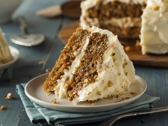 Ein Stück Karotten-Nuss-Kuchen mit Frischkäse auf  weißem Teller auf blauem Untergrund. | © Getty Images/ bhofack2