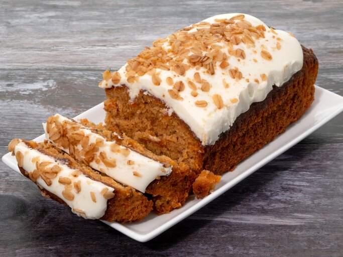 Karottenkuchen mit Creme drapiert auf weißer Platte auf grauem Holz. | © Getty Images/clubfoto