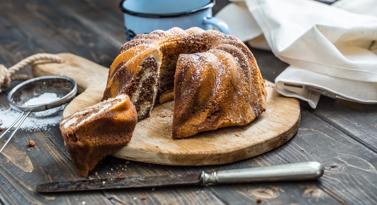 Ein frisch gebackener Marmorkuchen in Gugelhupfform, bestäubt mit Puderzucker, präsentiert auf einem Holzbrett. 