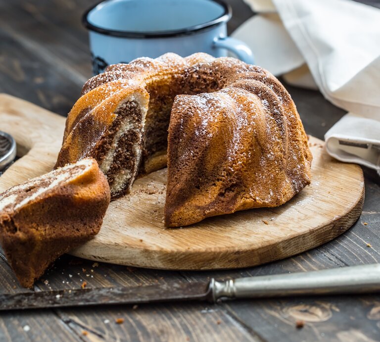 Ein frisch gebackener Marmorkuchen in Gugelhupfform, bestäubt mit Puderzucker, präsentiert auf einem Holzbrett.  | © GettyImages/MarianVejcik