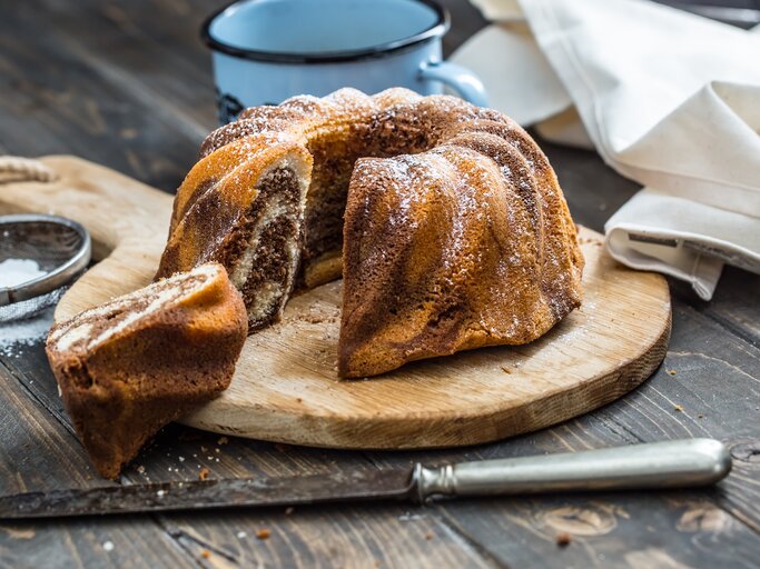 Ein frisch gebackener Marmorkuchen in Gugelhupfform, bestäubt mit Puderzucker, präsentiert auf einem Holzbrett.  | © GettyImages/MarianVejcik