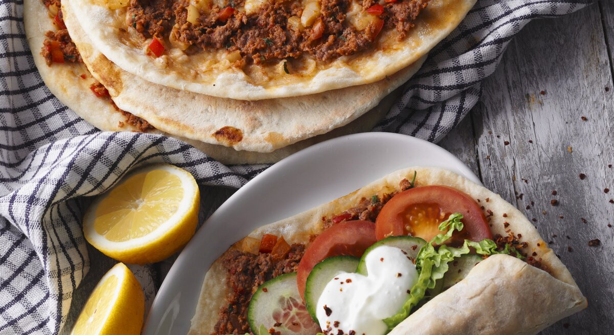 Draufsicht: Vier türkische Pizza Lahmacun. Drei nur mit Hackfleisch, eine mit Tomaten, Gurkenscheiben, Salat und Joghurtsauce