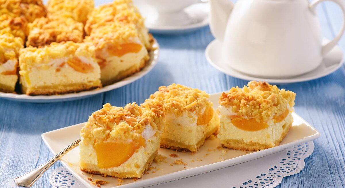 Geschnittener Pfirsich-Käsekuchen mit Streusel  auf weißem Geschirr auf blauer, hölzerner Oberfläche.