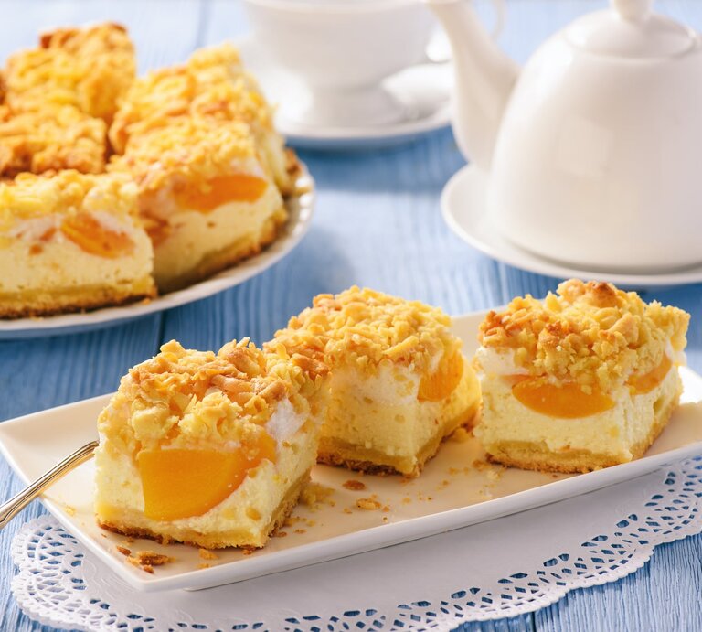 Geschnittener Pfirsich-Käsekuchen mit Streusel  auf weißem Geschirr auf blauer, hölzerner Oberfläche. | © Getty Images/iko636