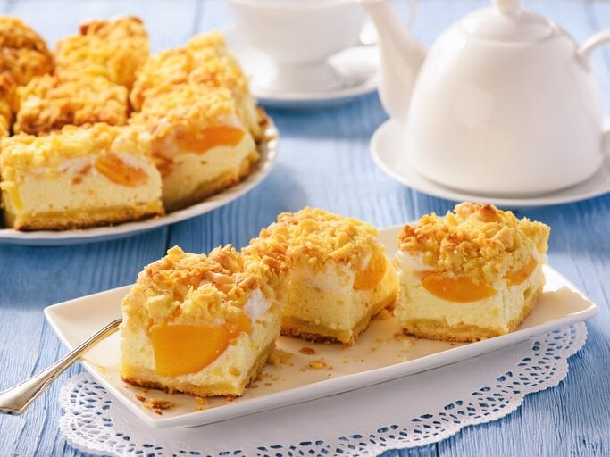 Geschnittener Pfirsich-Käsekuchen mit Streusel  auf weißem Geschirr auf blauer, hölzerner Oberfläche. | © Getty Images/iko636