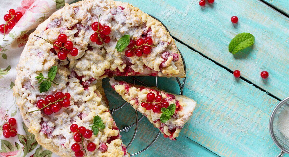 Johannisbeerkuchen mit Streusel auf einem Kuchengitter vor blauem Hintergrund. Links eine Tischdecke.
