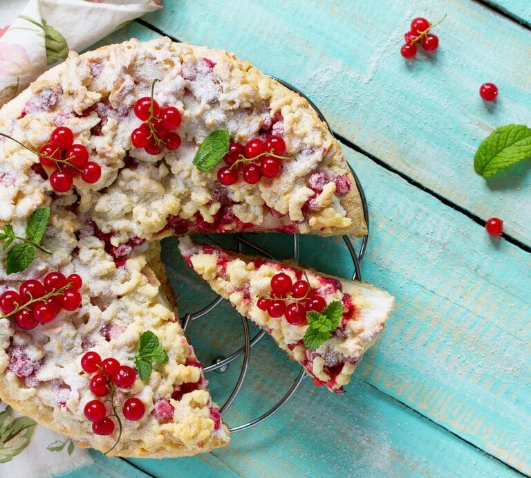 Johannisbeerkuchen mit Streusel auf einem Kuchengitter vor blauem Hintergrund. Links eine Tischdecke. | © iStock/as117pxz