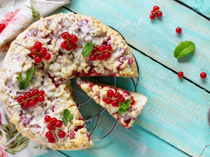 Johannisbeerkuchen mit Streusel auf einem Kuchengitter vor blauem Hintergrund. Links eine Tischdecke. | © iStock/as117pxz