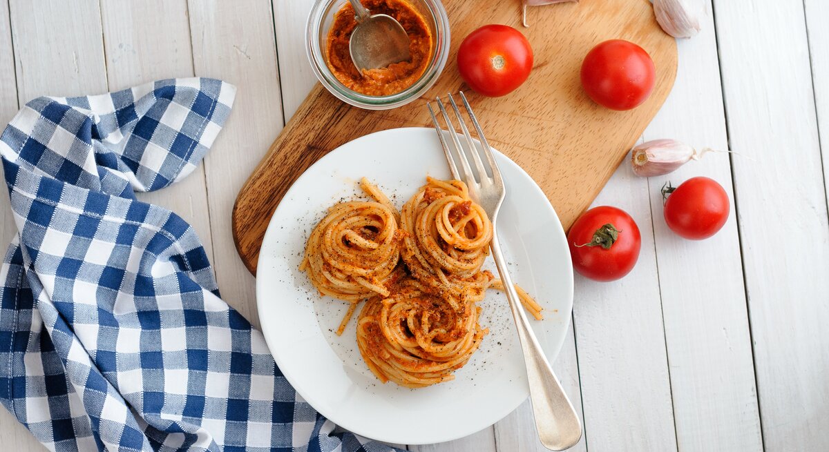 Aufgedrehte Spaghetti-Nester mit Pesto rosso auf weißem Teller. Daneben Tomaten und Knoblauch. 