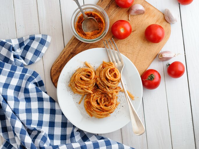 Aufgedrehte Spaghetti-Nester mit Pesto rosso auf weißem Teller. Daneben Tomaten und Knoblauch.  | © iStock/instamatics
