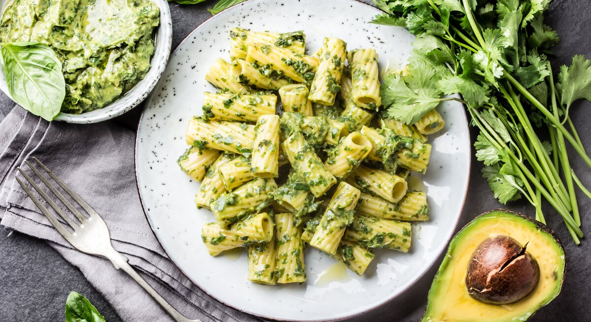 Weißer teller, darauf angerichtet Penne mit einem cremigen Avocado-Pesto. Die Zutaten sind drumherum verteilt. 