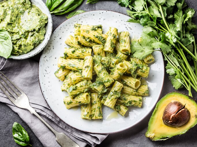 Weißer teller, darauf angerichtet Penne mit einem cremigen Avocado-Pesto. Die Zutaten sind drumherum verteilt.  | © iStock/LarisaBlinova