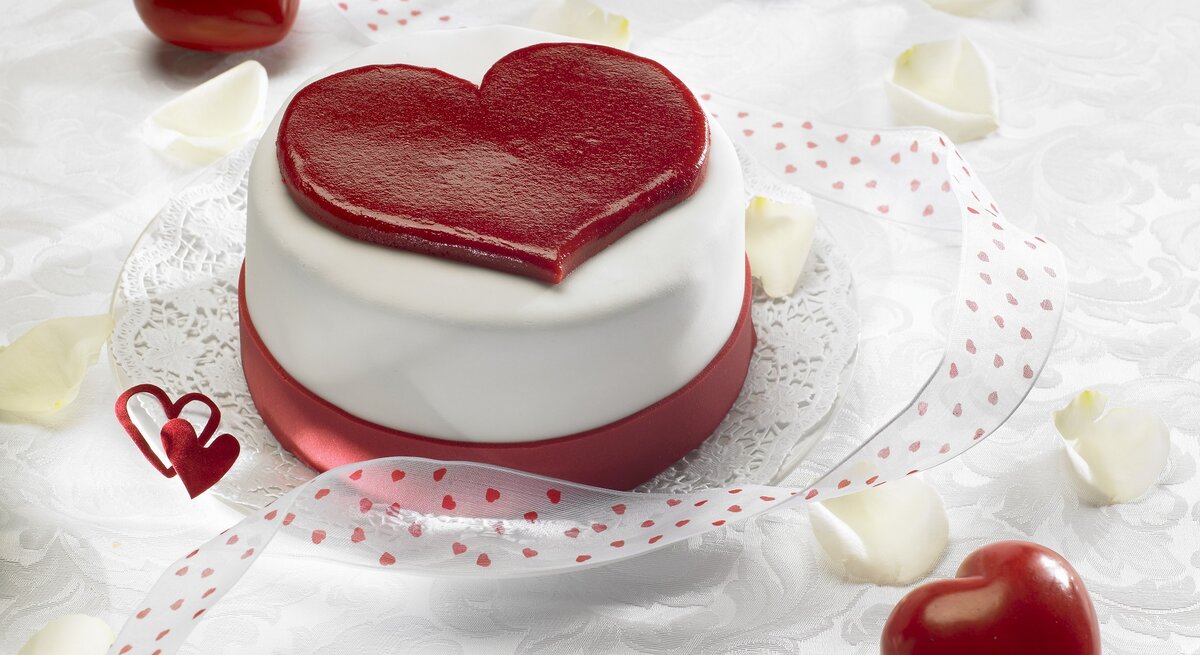 Ein herzförmiger Kuchen mit glatter, weißer Glasur und einem großen, roten Herz auf der Oberseite, der auf einem Tisch mit weißer Spitzenunterlage und roten Herzen verziert ist. In der Umgebung sind rote Herzen und weiße Blütenblätter verstreut, zusammen mit einem roten Band mit Herzmuster, das den Kuchen umgibt. Das Bild strahlt eine romantische und festliche Stimmung aus, ideal für Valentinstag oder einen besonderen Anlass.