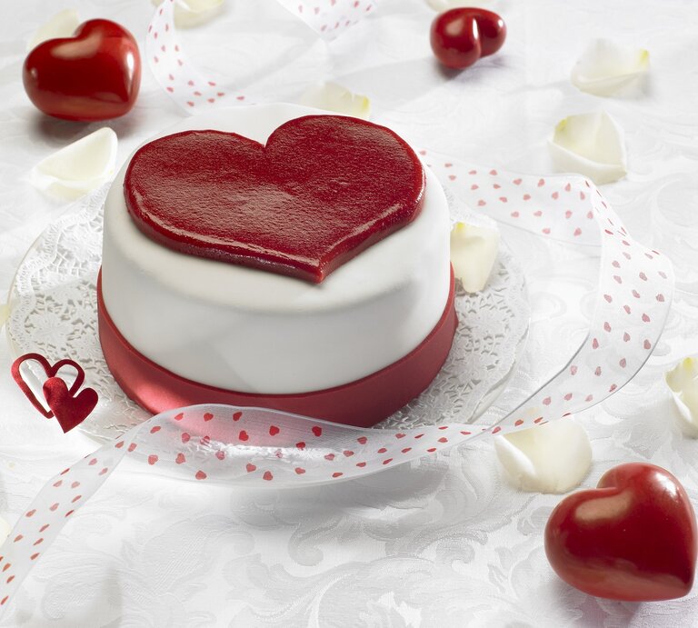 Ein herzförmiger Kuchen mit glatter, weißer Glasur und einem großen, roten Herz auf der Oberseite, der auf einem Tisch mit weißer Spitzenunterlage und roten Herzen verziert ist. In der Umgebung sind rote Herzen und weiße Blütenblätter verstreut, zusammen mit einem roten Band mit Herzmuster, das den Kuchen umgibt. Das Bild strahlt eine romantische und festliche Stimmung aus, ideal für Valentinstag oder einen besonderen Anlass. | © StockFood / Schmidt, Petra