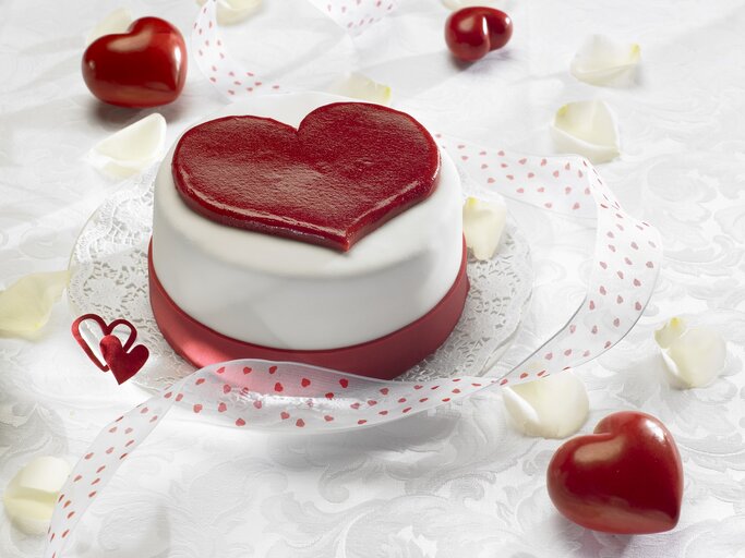 Ein herzförmiger Kuchen mit glatter, weißer Glasur und einem großen, roten Herz auf der Oberseite, der auf einem Tisch mit weißer Spitzenunterlage und roten Herzen verziert ist. In der Umgebung sind rote Herzen und weiße Blütenblätter verstreut, zusammen mit einem roten Band mit Herzmuster, das den Kuchen umgibt. Das Bild strahlt eine romantische und festliche Stimmung aus, ideal für Valentinstag oder einen besonderen Anlass. | © StockFood / Schmidt, Petra