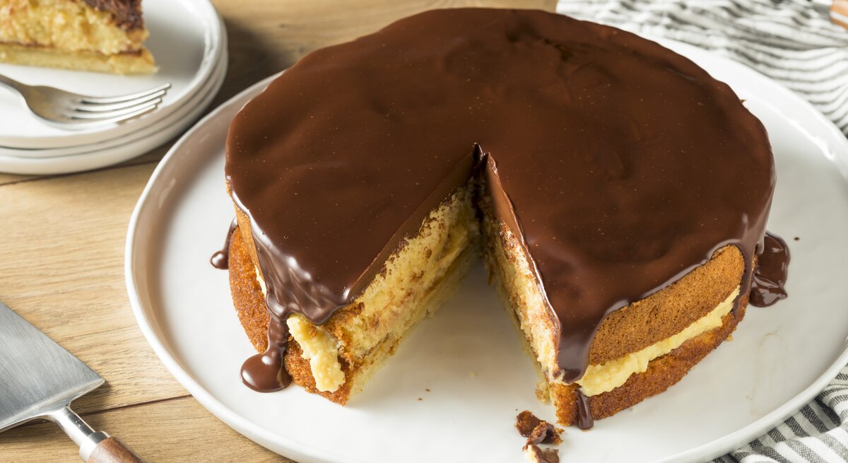 Ein Bild von einem Boston Cream Pie auf einem weißen Teller, mit einem Stück herausgeschnitten, zeigt die Schichten aus fluffigem Kuchen, cremiger Vanillefüllung und glänzender Schokoladenglasur, serviert auf einem Holztisch mit einem gestreiften Küchentuch im Hintergrund.