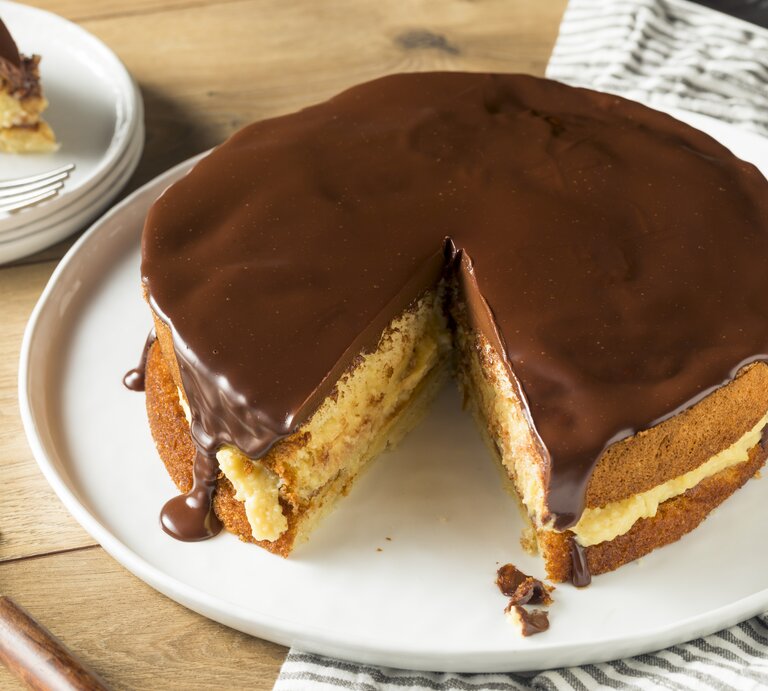 Ein Bild von einem Boston Cream Pie auf einem weißen Teller, mit einem Stück herausgeschnitten, zeigt die Schichten aus fluffigem Kuchen, cremiger Vanillefüllung und glänzender Schokoladenglasur, serviert auf einem Holztisch mit einem gestreiften Küchentuch im Hintergrund. | © iStock.com/bhofack2