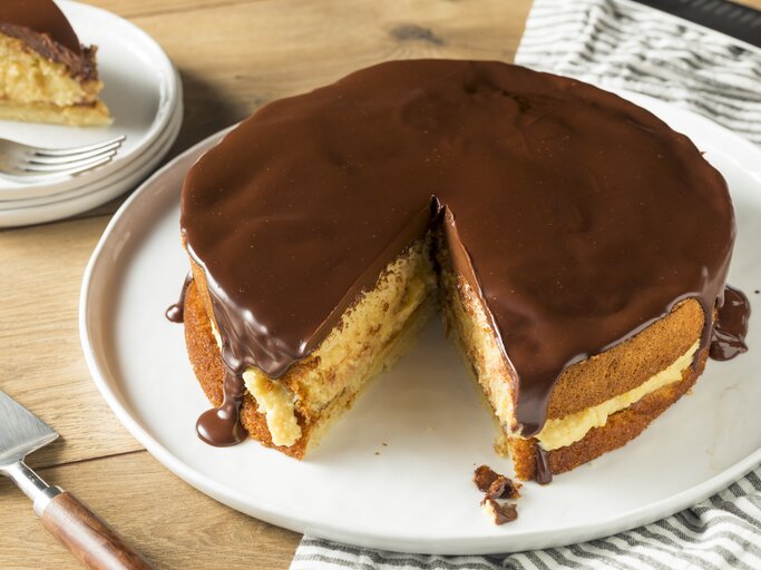 Ein Bild von einem Boston Cream Pie auf einem weißen Teller, mit einem Stück herausgeschnitten, zeigt die Schichten aus fluffigem Kuchen, cremiger Vanillefüllung und glänzender Schokoladenglasur, serviert auf einem Holztisch mit einem gestreiften Küchentuch im Hintergrund. | © iStock.com/bhofack2